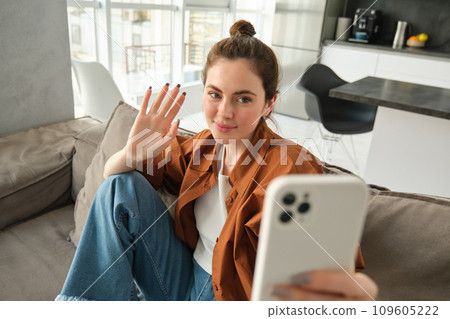Happy woman is sitting at home with smartphone, online chatting, waving hand at mobile phone, saying hello, talking to friends using application, sitting on sofa at home 109605222