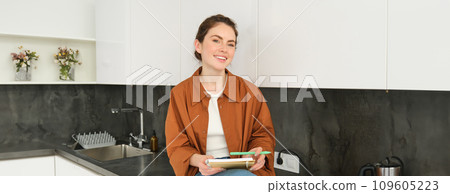 Portrait of happy cute woman at home, writing in notebook, makes notes in her diary, has a planner in her hands, sits in kitchen Portrait of happy cute woman at home, writing in notebook, makes notes in her diary, has a planner in her hands, sits in kitchen 109605223
