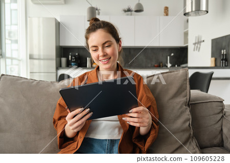 Portrait of young brunette woman reading on digital tablet, watching tv series on her application, sitting on couch in living room Portrait of young brunette woman reading on digital tablet, watching tv series on her application, sitting on couch in living room 109605228