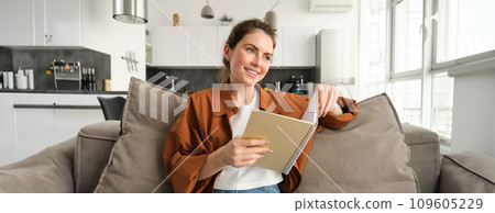 Smiling young brunette woman, sits on sofa in living room, holds notebook, reads her notes, studies for exam, student does her homework at home Smiling young brunette woman, sits on sofa in living room, holds notebook, reads her notes, studies for exam, student does her homework at home 109605229