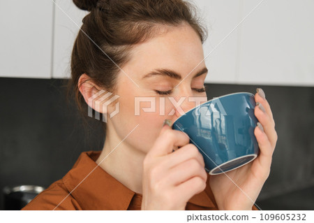 Portrait of beautiful young woman, enjoying delicious aroma of freshly made coffee, holding mug, standing near machine in the kitchen, drinking from mug Portrait of beautiful young woman, enjoying delicious aroma of freshly made coffee, holding mug, standing near machine in the kitchen, drinking from mug 109605232
