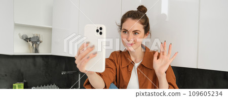 Portrait of young cheerful woman laughing and smiling during phone call, video chats with friend, sits on kitchen counter and talks to someone using smartphone app Portrait of young cheerful woman laughing and smiling during phone call, video chats with friend, sits on kitchen counter and talks to someone using smartphone app 109605234