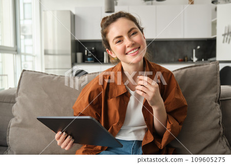 Portrait of young brunette woman reading on digital tablet, watching tv series on her application, sitting on couch in living room 109605275