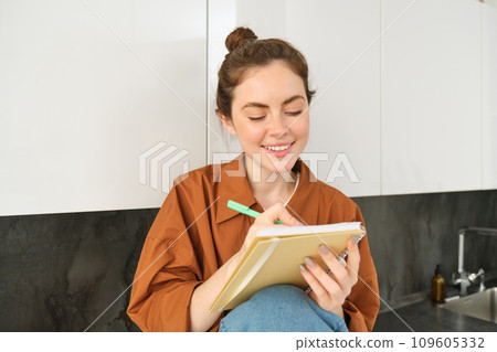 Portrait of young creative girl, writing down her ideas in planner, does homework in notebook, sits at home on kitchen counter 109605332