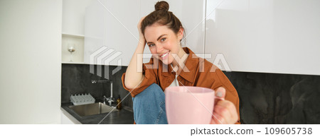 Pretty young smiling woman sits in kitchen, offers you cup of coffee, extends hand with pink mug, sharing her drink with someone Pretty young smiling woman sits in kitchen, offers you cup of coffee, extends hand with pink mug, sharing her drink with someone 109605738