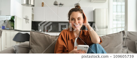 Beautiful smiling female model sitting on couch with smartphone, looking happy at camera, scrolling social media, messaging someone, doing online shopping Beautiful smiling female model sitting on couch with smartphone, looking happy at camera, scrolling social media, messaging someone, doing online shopping 109605739