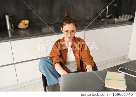 Portrait of beautiful young woman studying at home, sitting with laptop and documents in kitchen. Self-employed business owner typing on computer, sending emails to clients Portrait of beautiful young woman studying at home, sitting with laptop and documents in kitchen. Self-employed business owner typing on computer, sending emails to clients 109605740