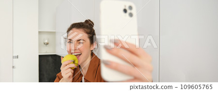 Portrait of happy, smiling young woman records herself, takes selfie while eating an apple in the kitchen, using smartphone app, makes photos with mobile phone 109605765
