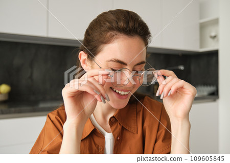 Portrait of young happy woman, sitting in kitchen, wearing glasses, reading or working from home 109605845