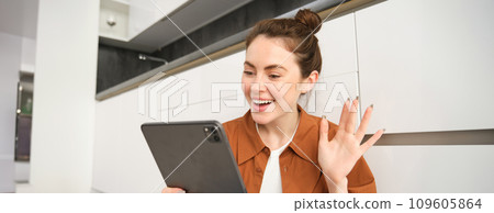 Portrait of friendly smiling woman video chats, connects to online conversation on digital tablet, says hello at camera Portrait of friendly smiling woman video chats, connects to online conversation on digital tablet, says hello at camera 109605864