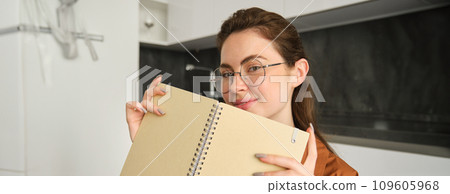 Close up portrait of smiling beautiful woman with notebook, making notes, writing in diary, studying at home, preparing homework at in kitchen Close up portrait of smiling beautiful woman with notebook, making notes, writing in diary, studying at home, preparing homework at in kitchen 109605968