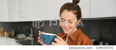 Portrait of beautiful young woman, enjoying delicious aroma of freshly made coffee, holding mug, standing near machine in the kitchen, drinking from mug 109605970