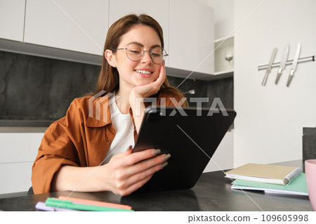 E-learning and remote workplace concept. Young woman in glasses, student studying at home in kitchen, reading on digital tablet, looking at her project, connects to online meeting 109605998