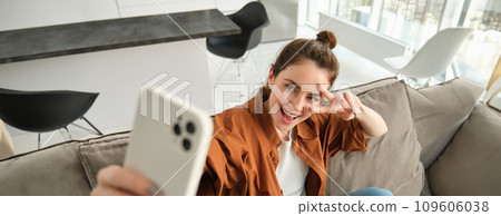 Positive and happy young woman taking selfie on smartphone, sitting on couch, showing peace, v-sign gesture Positive and happy young woman taking selfie on smartphone, sitting on couch, showing peace, v-sign gesture 109606038