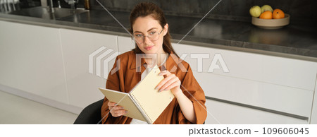 Portrait of young self-employed freelance tutor, sitting in kitchen at home, holding notebook with study notes, revising for university 109606045