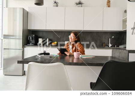 Portrait of young woman studying at home. Girl reading something on tablet, sitting in kitchen in glasses Portrait of young woman studying at home. Girl reading something on tablet, sitting in kitchen in glasses 109606084