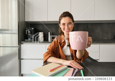Stylish young woman, student studying from home, sitting in kitchen with homework, giving you cup of coffee, raising pink mug, smiling at camera Stylish young woman, student studying from home, sitting in kitchen with homework, giving you cup of coffee, raising pink mug, smiling at camera 109606153