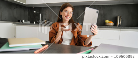 Portrait of young woman working from home, sitting in kitchen and talking with team, has online meeting, using digital tablet to join webinar, having conversation 109606892
