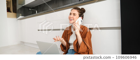 Cheerful smiling woman sits on floor in the kitchen with laptop, makes a phone call, talks to friend and laughs during conversation 109606946