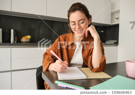 Portrait of modern young woman in glasses, student sitting at home and doing homework, writing down information in notebook and smiling, working on project Portrait of modern young woman in glasses, student sitting at home and doing homework, writing down information in notebook and smiling, working on project 109606963
