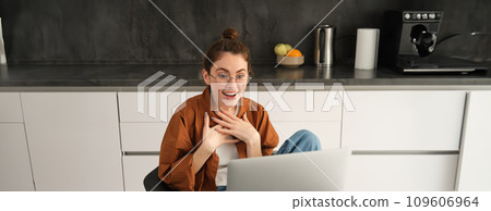 Portrait of beautiful woman in glasses, sits at home in kitchen, looks at computer screen with amazed, surprised face, reacts to big great news seeing it on laptop Portrait of beautiful woman in glasses, sits at home in kitchen, looks at computer screen with amazed, surprised face, reacts to big great news seeing it on laptop 109606964