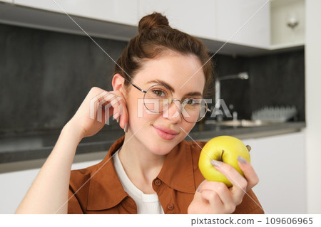 Beautiful young woman in glasses, sitting on chair in the kitchen, eating green apple and smiling Beautiful young woman in glasses, sitting on chair in the kitchen, eating green apple and smiling 109606965