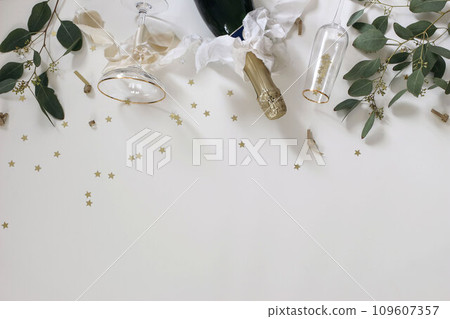 New year party, celebration. Bottle of champagne wine, drinking glasses, bows. Eucalyptus tree branches isolated on white table background. Golden star confetti decor. Birthday, anniversary. Flat lay 109607357
