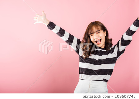 An Asian woman smiles, making a triumphant gesture with raised fists, celebrating her success. Studio shot on a pink background, capturing the joy and happiness of a winner. 109607745