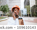 Happy black woman in helmet standing with coffee cup and speaking on smartphone 109610143