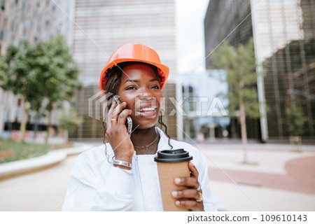 Happy black woman in helmet standing with coffee cup and speaking on smartphone Happy black woman in helmet standing with coffee cup and speaking on smartphone 109610143