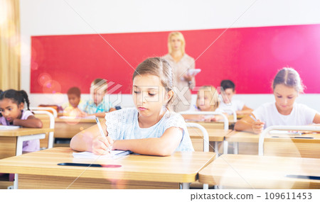 Preteen girl listening to teacher and writing in notebook in class 109611453