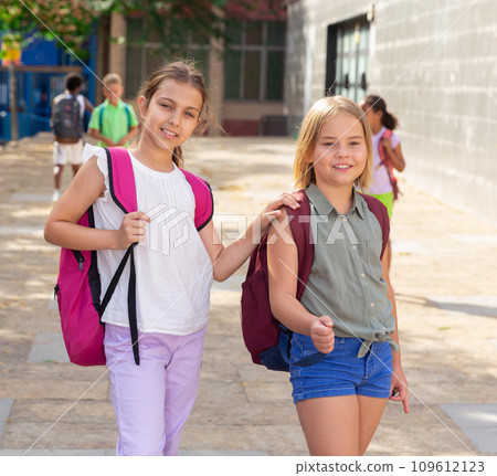 Friendly preteen girls with backpacks walking through city street 109612123
