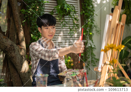A focused and handsome young Asian man is measuring his drawing on a canvas with a paintbrush. A focused and handsome young Asian man is measuring his drawing on a canvas with a paintbrush. 109613713