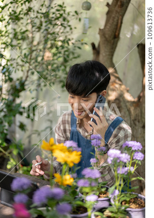 Young happy Asian male florist or small flower garden owner is talking on the phone with a supplier. Young happy Asian male florist or small flower garden owner is talking on the phone with a supplier. 109613736