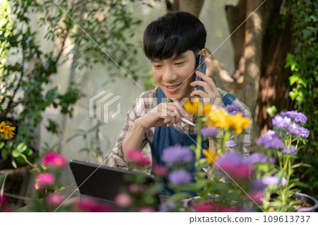Young happy Asian male florist or small flower garden owner is talking on the phone with a supplier. Young happy Asian male florist or small flower garden owner is talking on the phone with a supplier. 109613737