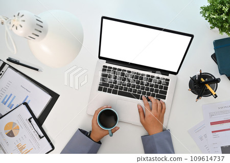 Female worker holding cup of coffee and using laptop on white office desk. Top view Female worker holding cup of coffee and using laptop on white office desk. Top view 109614737