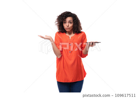 young european woman with curly black hair in an orange blouse shrugs her shoulders holding a phone young european woman with curly black hair in an orange blouse shrugs her shoulders holding a phone 109617511