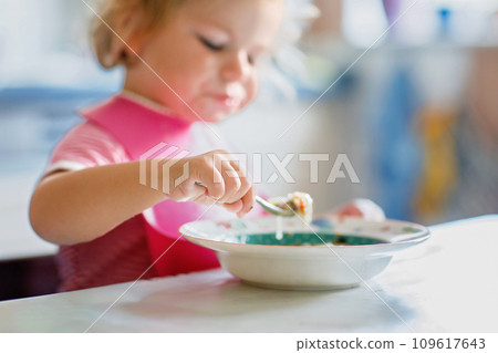 Adorable baby girl eating from spoon vegetable noodle soup. Healthy food, child, feeding and development concept. Cute toddler child with spoon sitting in highchair and learning to eat by itself 109617643