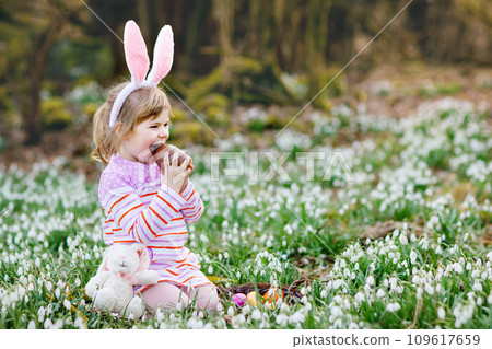 Little girl with Easter bunny ears making egg hunt in spring forest on sunny day, outdoors. Cute happy child with lots of snowdrop flowers, huge chocolate egg and colored eggs. 109617659