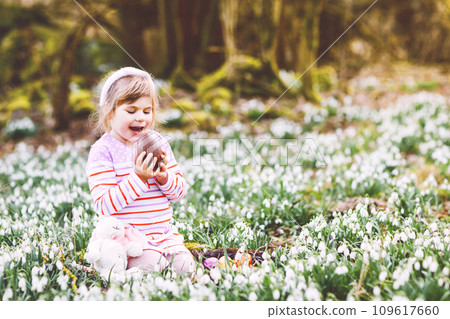 Little girl with Easter bunny ears making egg hunt in spring forest on sunny day, outdoors. Cute happy child with lots of snowdrop flowers, huge chocolate egg and colored eggs. Little girl with Easter bunny ears making egg hunt in spring forest on sunny day, outdoors. Cute happy child with lots of snowdrop flowers, huge chocolate egg and colored eggs. 109617660