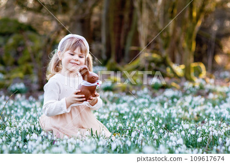 Happy little girl with Easter bunny ears eating chocolate figure in spring forest on sunny day, outdoors. Cute child with lots of snowdrop flowers. Springtime, christian holiday concept. 109617674