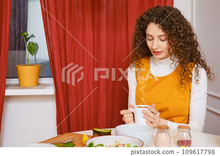 Peaceful woman holds cup of coffee with both hands while having breakfast 109617798
