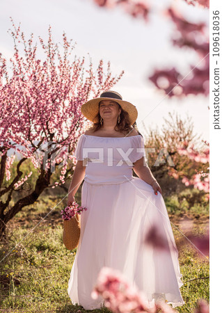 Woman blooming peach orchard. Against the backdrop of a picturesque peach orchard, a woman in a long white dress and hat enjoys a peaceful walk in the park, surrounded by the beauty of nature. Woman blooming peach orchard. Against the backdrop of a picturesque peach orchard, a woman in a long white dress and hat enjoys a peaceful walk in the park, surrounded by the beauty of nature. 109618036