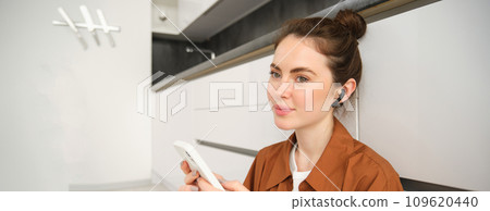 Carefree smiling woman with smartphone, messaging, using mobile phone, sitting on kitchen floor, listening music in wireless headphones 109620440