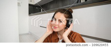 Portrait of beautiful female model on kitchen floor at home, listens music in wireless headphones, smiling and looking happy 109620455
