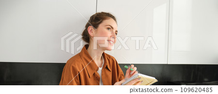 Portrait of woman writing down her ideas in a notebook, holding pen and looking thoughtful, inspired to write in diary, plans her schedule or makes a list for grocery shopping 109620485
