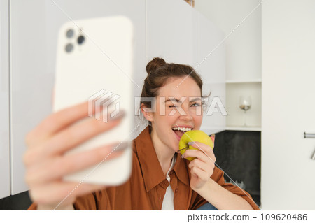 Portrait of happy, smiling young woman records herself, takes selfie while eating an apple in the kitchen, using smartphone app, makes photos with mobile phone 109620486