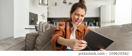 Portrait of young woman playing video games on digital tablet, looking excited, tilting gadget and looking at screen, sitting on couch in living room 109620562