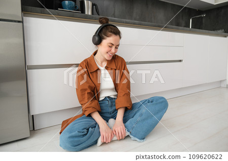 Cute modern girl on kitchen floor at home, listening music in wireless headphones, having fun 109620622