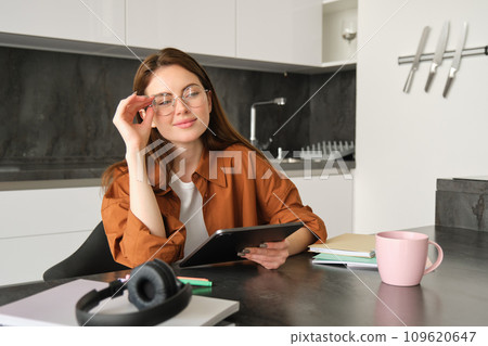 Portrait of young student, woman studying at home, working remotely, setup workplace in her kitchen, sitting on chair with digital tablet, reading in glasses 109620647